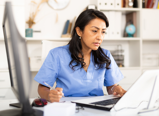 A woman wearing blue scrubs is sitting at a desk, focused on her laptop.