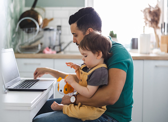 A man is sitting at a desk using a laptop while holding a young child in his lap.