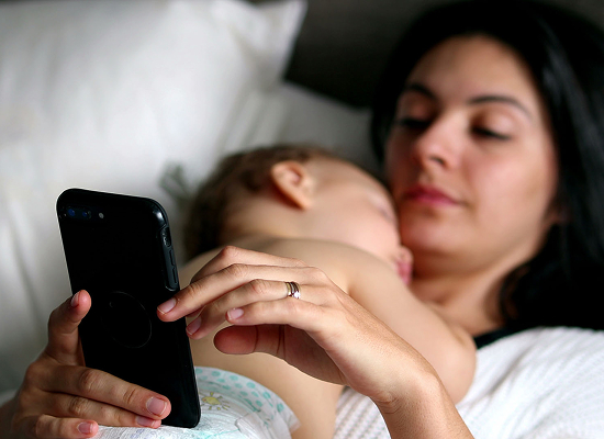 A woman is lying down in bed, holding a smartphone while cradling a sleeping baby against her chest.
