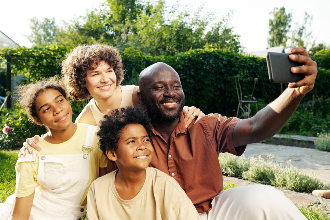 Young cheerful family of father, mother and two kids taking selfie together while relaxing on green lawn not far from their country house