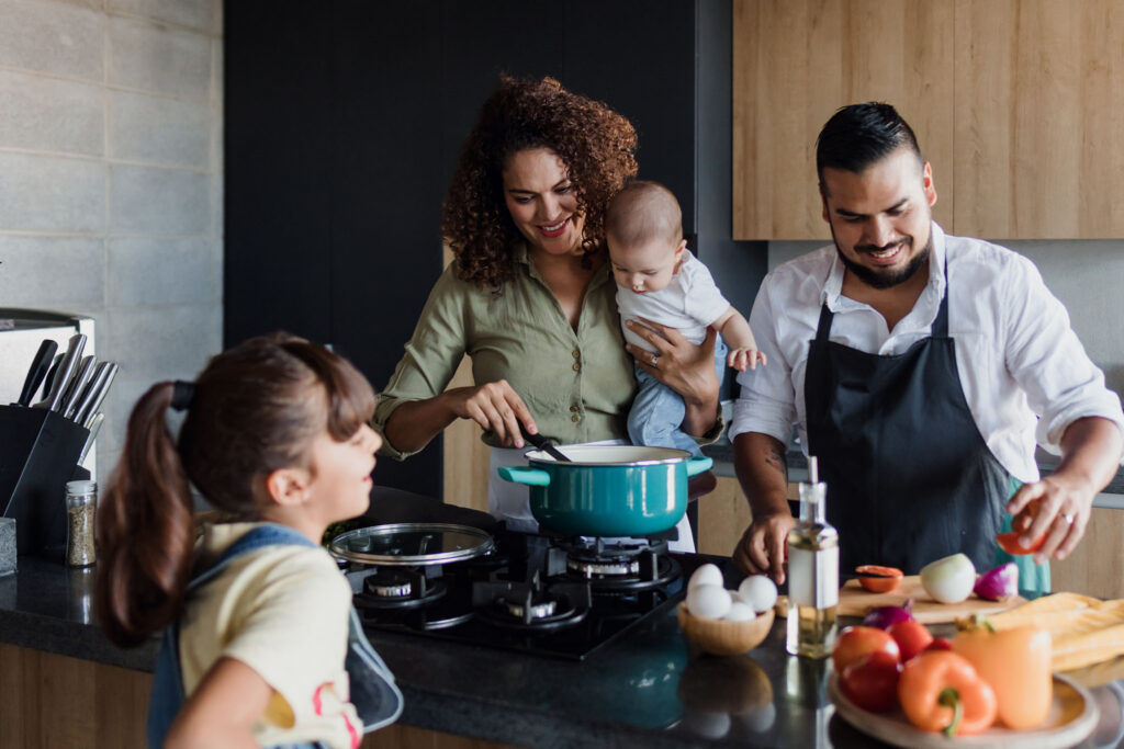 A family is cooking in a modern kitchen with vegetables, eggs, and olive oil on the countertop. Parents prepare a meal as their daughter looks on.