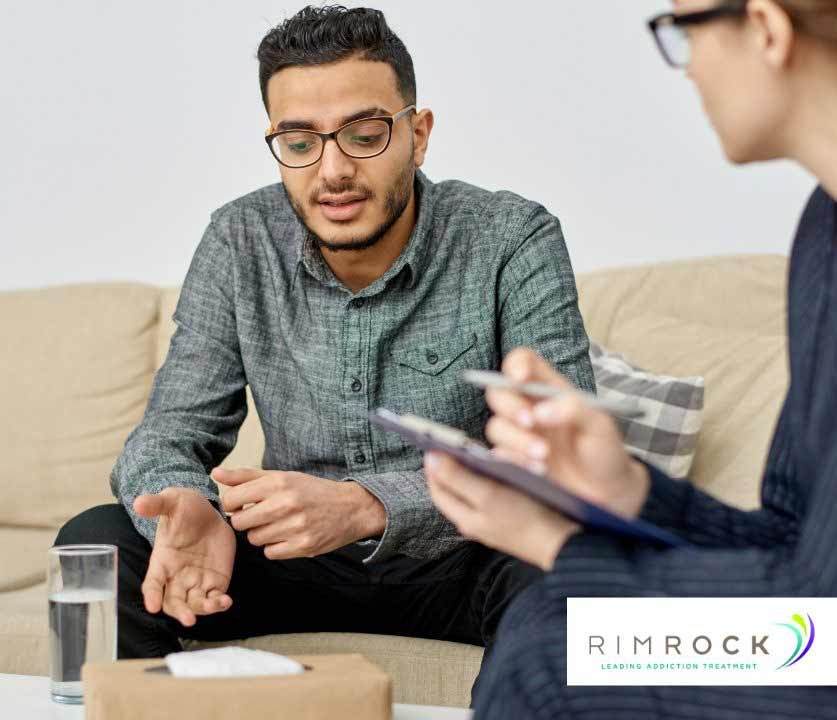 A man is sitting on a couch, speaking with a woman who is taking notes on a clipboard.
